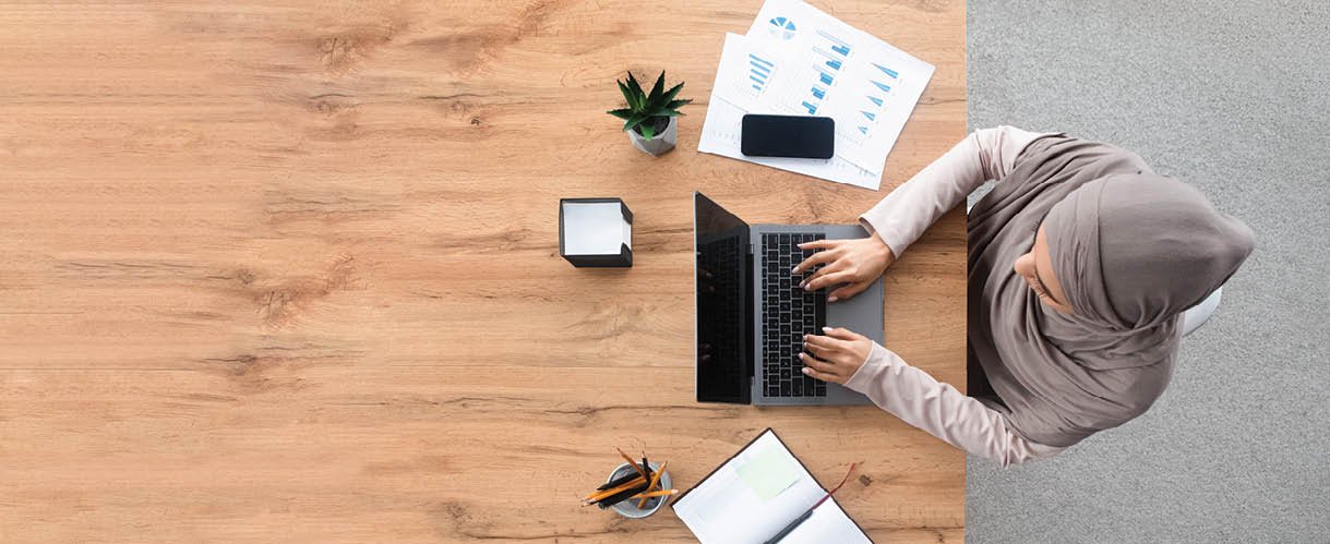 Long shot of muslim female employee in hijab working on laptop computer at workplace in modern office, horizontal banner with free space, top view
